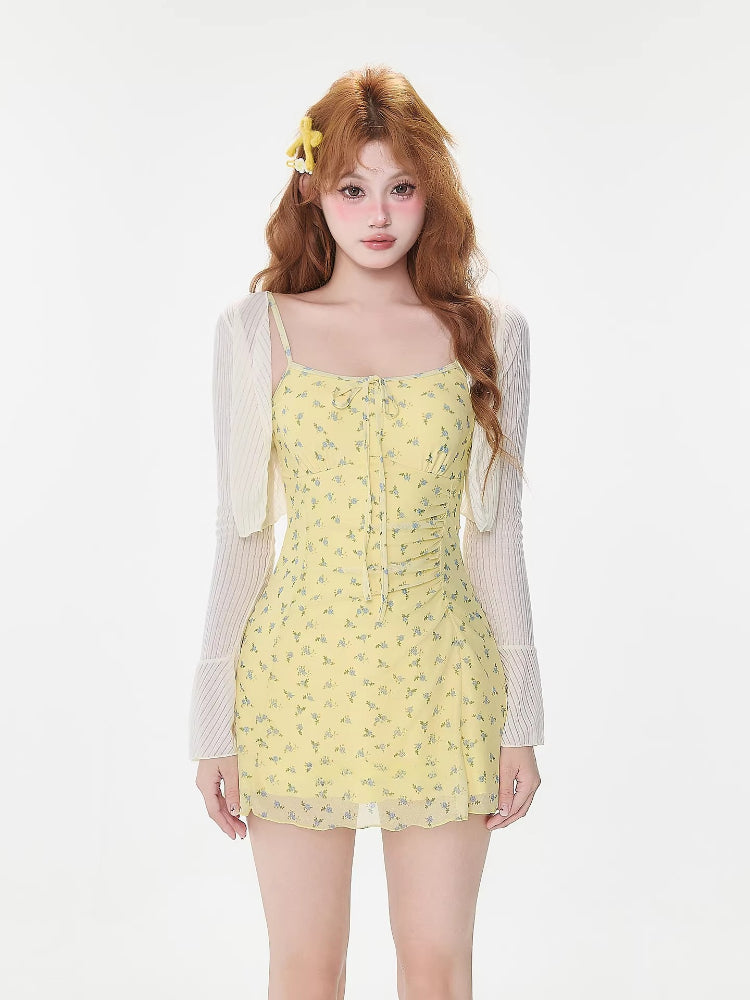 Woman wearing a yellow floral print mini dress with spaghetti straps and a tie front detail, paired with a white sheer long-sleeve shrug.