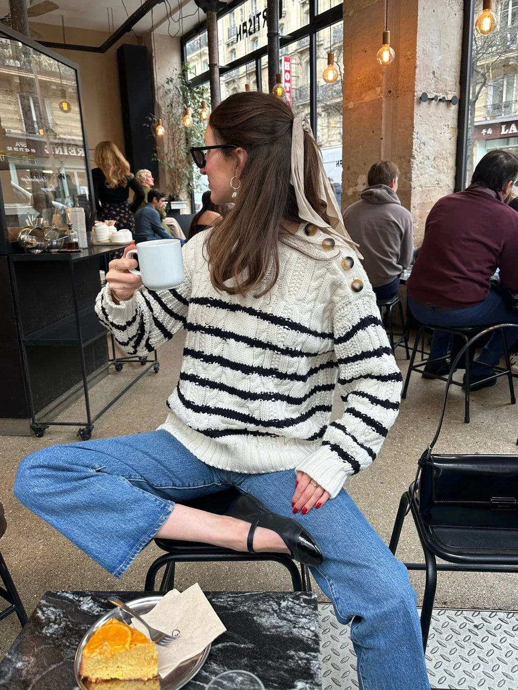 Woman in a striped knit sweater and blue jeans holding a coffee cup while sitting in a cafe.
