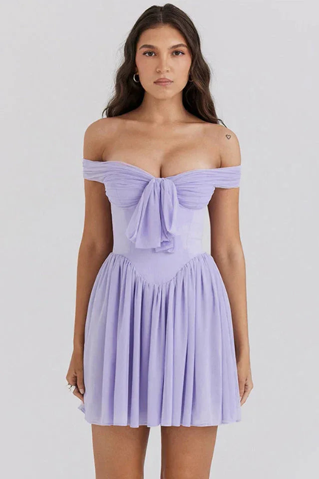 Woman wearing a light purple off-the-shoulder mini dress with a bow detail at the bust and a pleated skirt, front view.