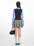 Back view of a woman wearing a blue striped collared shirt, navy V-neck vest, and grey pleated skirt.