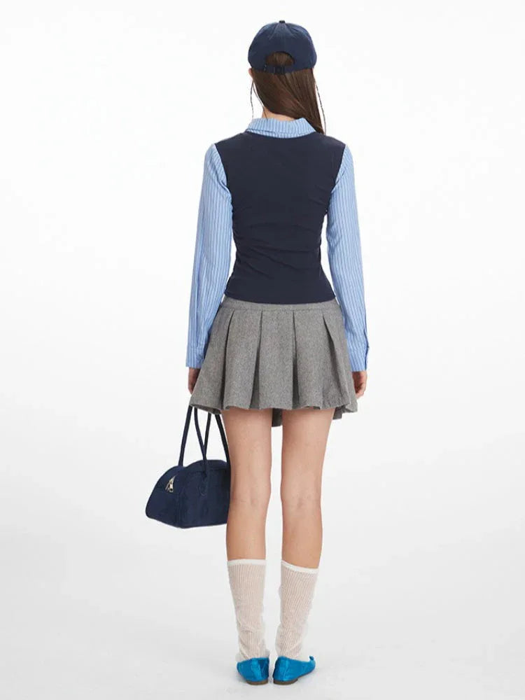 Back view of a woman wearing a blue striped collared shirt, navy V-neck vest, and grey pleated skirt.