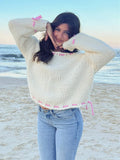 Woman posing on a beach wearing a cream knit sweater with pink stitching and ties, paired with denim jeans.