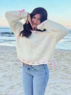 Woman posing on a beach wearing a cream knit sweater with pink stitching and ties, paired with denim jeans.