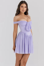 Woman wearing a light purple off-the-shoulder mini dress with a bow detail at the bust and a pleated skirt, three-quarter front view.