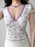Close-up of a woman wearing a white mesh top with a purple floral print and purple lace trim around the V-neckline. The top features a tie closure at the bust and short sleeves.