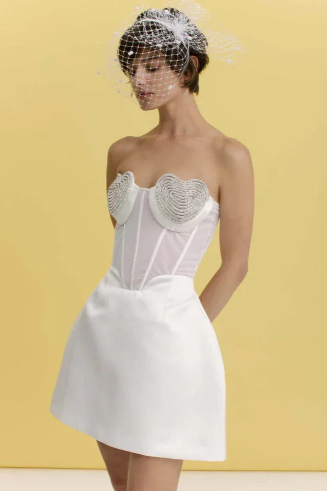 Front view of a model in a white strapless mini dress with a corset bodice decorated with heart-shaped embellishments, shown with a yellow backdrop.