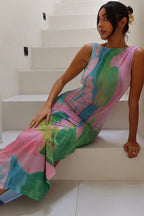 Woman wearing a long, sleeveless pink, green, and blue abstract print sheer mesh dress with a side slit, seated on a white staircase.