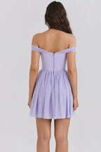 Woman wearing a light purple off-the-shoulder mini dress with a bow detail at the bust and a pleated skirt, back view.