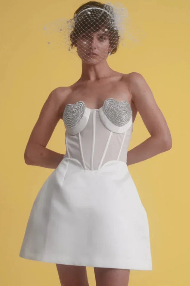 Model wearing a white strapless mini dress with a corset bodice featuring heart-shaped embellishments and a sheer panel, set against a yellow background.