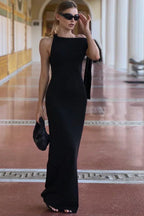 Woman wearing a black sleeveless column dress with an asymmetrical neckline and draped shoulder detail, holding a black handbag.