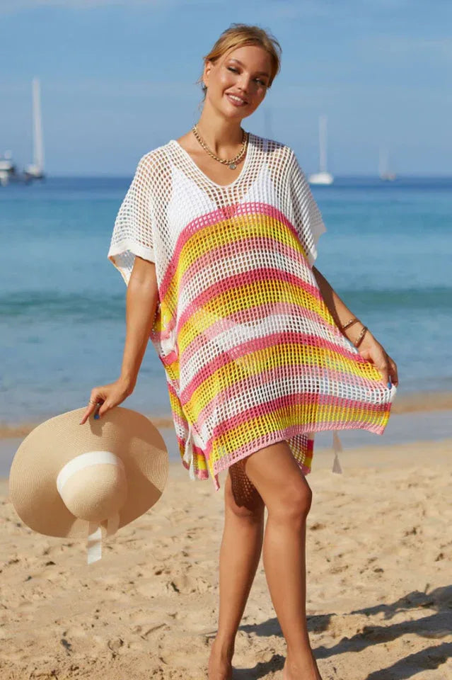 Woman holding a straw hat, wearing a white and striped mesh knit tunic on a beach.