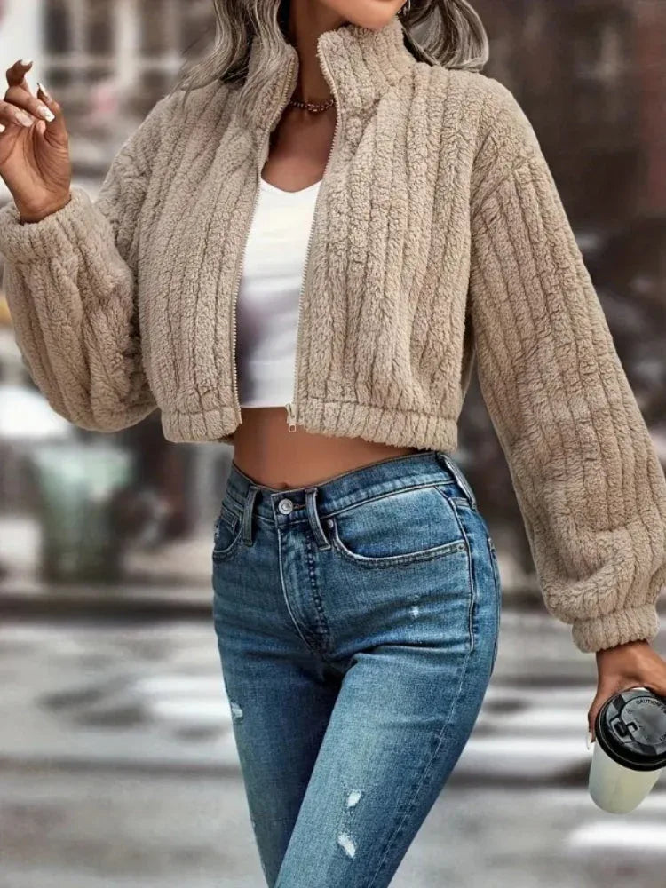 Woman in a tan fuzzy fleece cropped bomber jacket and blue jeans, holding a coffee cup.