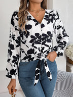 Woman wearing a white long-sleeve blouse with a black floral print and a tie-front detail, paired with blue denim jeans.