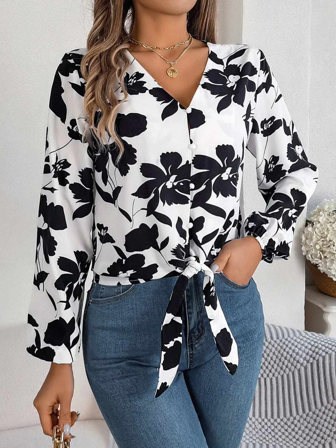 Woman wearing a white long-sleeve blouse with a black floral print and a tie-front detail, paired with blue denim jeans.