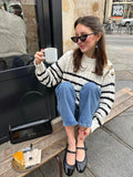 Woman wearing a striped knit sweater, blue jeans, and black ballet flats sitting on a bench outside a cafe.