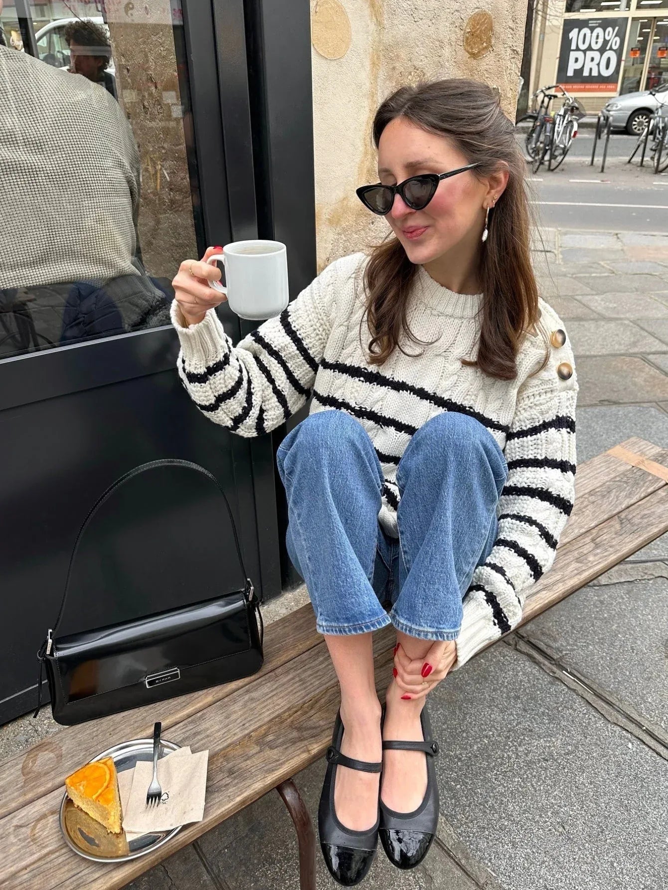 Woman wearing a striped knit sweater, blue jeans, and black ballet flats sitting on a bench outside a cafe.