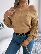 Woman posing in a tan off-the-shoulder cable knit cropped sweater, blue jeans, and holding a white handbag.