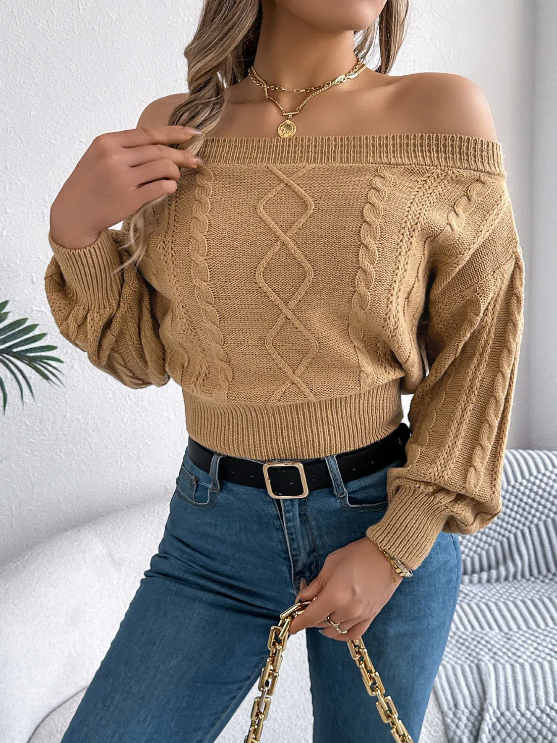 Woman posing in a tan off-the-shoulder cable knit cropped sweater, blue jeans, and holding a white handbag.