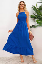 Woman wearing a royal blue smocked halter maxi dress with a tiered ruffle hem and crisscross back straps, holding a pink clutch.