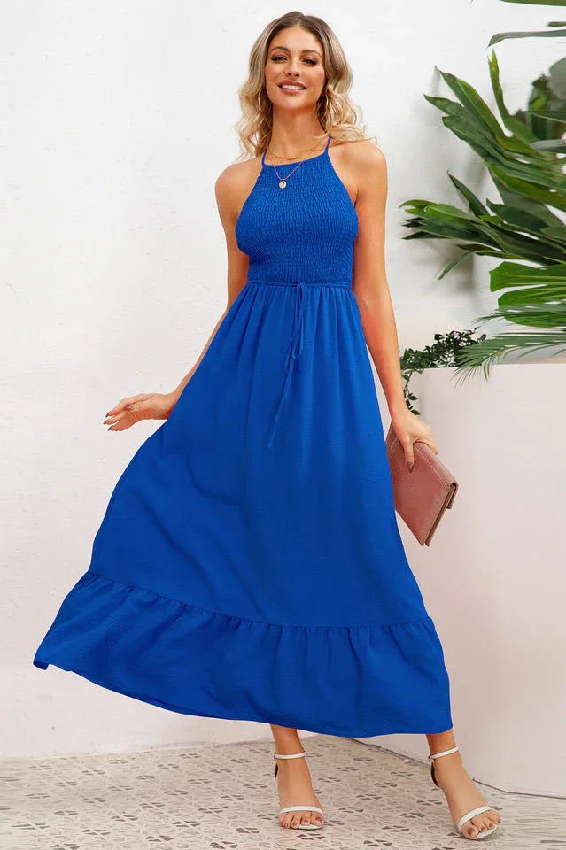Woman wearing a royal blue smocked halter maxi dress with a tiered ruffle hem and crisscross back straps, holding a pink clutch.