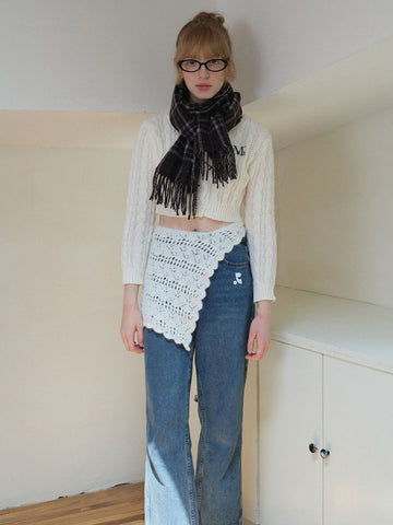 Woman wearing a cream cable knit cropped sweater with embroidered initial, a plaid scarf, and a white crocheted overlay skirt.
