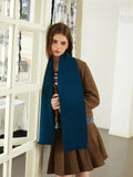 Woman leaning against a door frame wearing a brown tweed jacket, blue knit scarf, and brown pleated skirt.