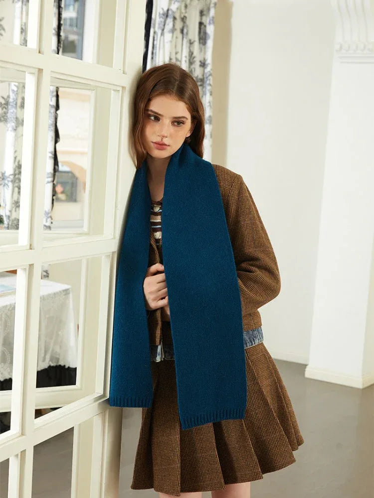 Woman leaning against a door frame wearing a brown tweed jacket, blue knit scarf, and brown pleated skirt.