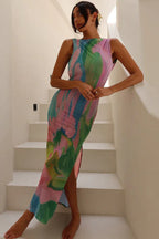 Woman wearing a long, sleeveless pink, green, and blue abstract print sheer mesh dress with a side slit, leaning against a white railing.