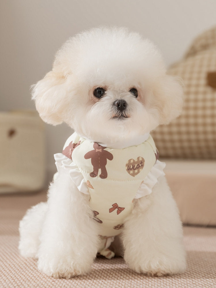 A Bichon Frise dog wears a light yellow padded vest with a teddy bear print and white ruffle trim.