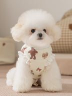 A Bichon Frise dog wears a light yellow padded vest with a teddy bear print and white ruffle trim.