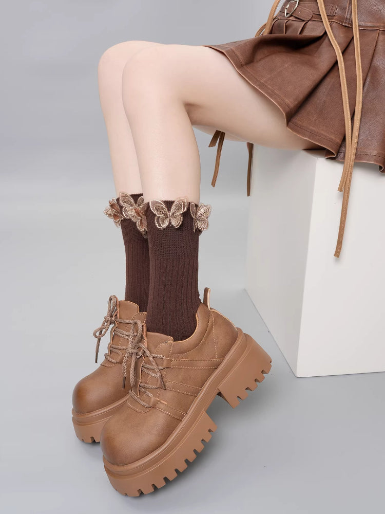 Close-up of brown ribbed crew socks featuring butterfly embellishments and a ruffled cuff, worn with brown platform shoes.