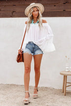 Model posing in a white off-the-shoulder top with voluminous bell sleeves, paired with distressed denim shorts.