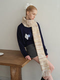 Model posing while leaning on a wooden table, wearing a navy argyle sweater, grey shorts, and a fringed scarf.