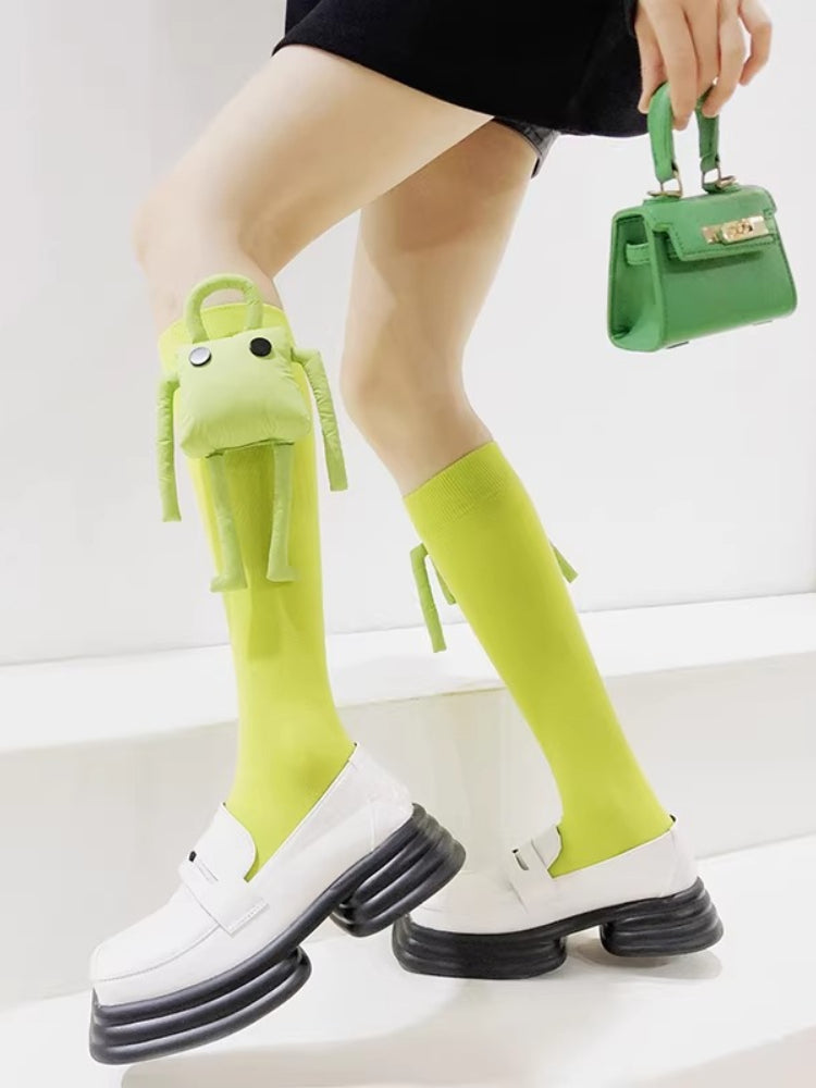 Close-up of lime green knee-high socks featuring a plush frog charm and tie detail, paired with white platform loafers.