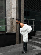 Woman walking on a cobblestone street wearing a cream faux fur cropped jacket and grey trousers.