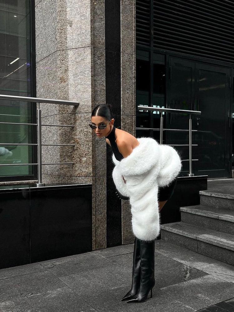 Woman walking on a cobblestone street wearing a cream faux fur cropped jacket and grey trousers.