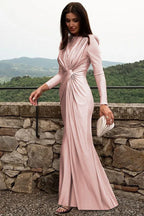 Woman in a long-sleeved, ruched pink satin gown walking outdoors with a clutch.