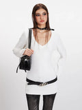 Front view of a woman in a white long-sleeve V-neck top, black belt, and holding a black handbag.