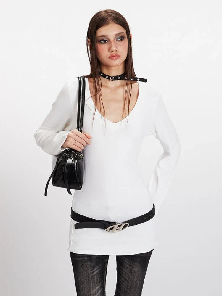 Front view of a woman in a white long-sleeve V-neck top, black belt, and holding a black handbag.