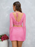 Woman wearing a pink sparkle mesh crop top with bell sleeves and a matching mini skirt, showing the back with a cowl neckline and tie closure.