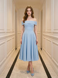 Woman wearing a light blue off-the-shoulder midi dress with a fitted bodice and full skirt, standing in a hallway.