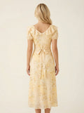 Back view of a woman in a yellow floral midi dress with a cinched waist and tie detail.