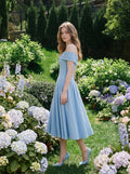 Woman in a light blue off-the-shoulder midi dress with a fitted bodice and full skirt, standing in a garden with flowers.