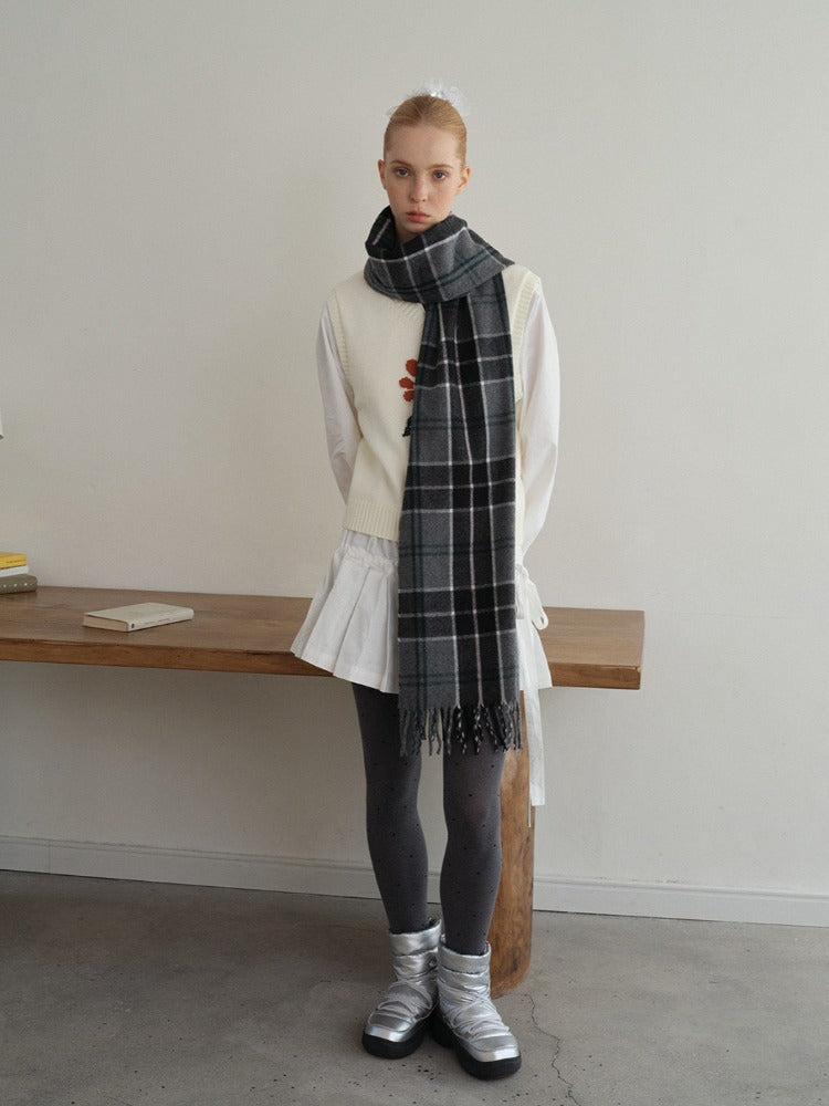 Full body shot of a woman in a white vest over a white long-sleeve shirt, a white pleated skirt, grey polka dot tights, and silver boots, with a dark plaid scarf.