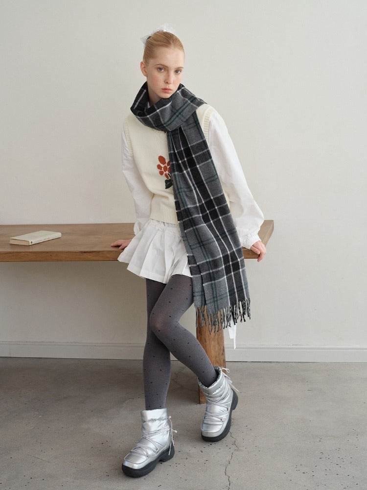 Model posing in a white vest, white shirt, white pleated skirt, grey polka dot tights, and silver boots, wearing a dark plaid scarf.