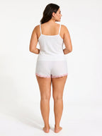 Woman wearing a white ribbed camisole and shorts pajama set with pink lace trim and bow details, back view.