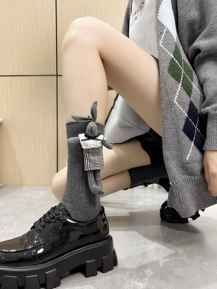Woman in grey ribbed knee-high socks with a plush bunny charm, kneeling next to black platform shoes.