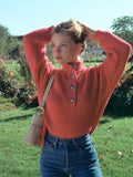 Woman wearing a coral knit sweater with a high neckline and button placket, paired with blue denim jeans, outdoors in a park.