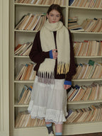 Woman posing with a cream scarf with ombre fringe in shades of yellow and purple, layered over a maroon sweater and white skirt.