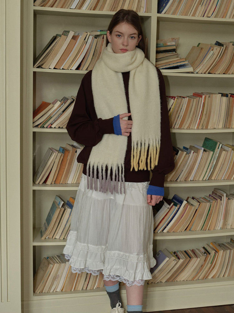 Woman posing with a cream scarf with ombre fringe in shades of yellow and purple, layered over a maroon sweater and white skirt.